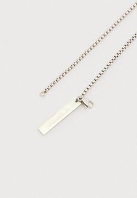 Ezüst lánc nyaklánc téglalap alakú medállal, amelyen az "FAVELA" felirat látható, és egy homárkapoccsal világos háttéren.