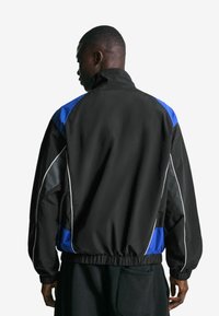 Man met een zwart-blauwe jas met witte piping en zwarte broek, kijkend in de richting van de achtergrond tegen een effen witte achtergrond.