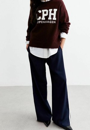 Personne portant un pull bordeaux « CPH Copenhagen » sur une chemise blanche, un pantalon large bleu marine, et des chaussures noires à bouts pointus, les mains dans les poches.