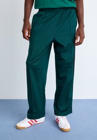 Grønne atletiske bukser lavet af letvægtsstof med elastisk talje, der har sidelapper, og de er parret med hvide sneakers med røde accenter.