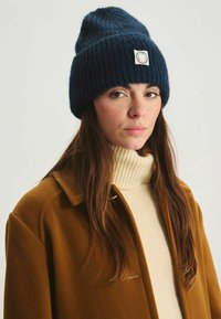 Bonnet tricoté bleu marine avec texture côtelée et patch logo, associé à un col roulé crème et un manteau marron.