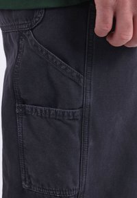 Pantalon en denim noir avec une poche latérale et des coutures visibles. Le tissu a une texture lisse avec une légère brillance.