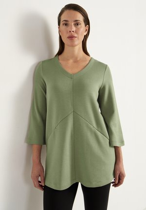 Grøn langærmet tunika med V-udskæring, der har et skråt sømdesign og en subtilt struktureret stof. Kombinationen med sorte leggings.