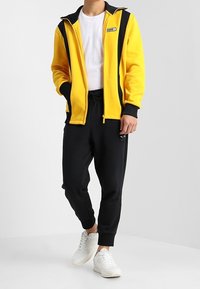 Gul fleecejacka med svarta ränder, dragkedjeslutning och logo; matchad med svarta joggingbyxor och vita sneakers. Slät textur, sportig design.