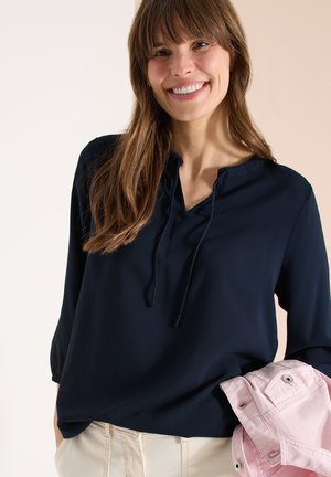 Femme aux longs cheveux bruns portant un chemisier bleu marine et un pantalon beige, tenant une veste rose pliée dans sa main gauche.