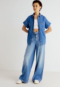 Jeanshemd in Mittelblau mit kurzen Ärmeln und Fronttaschen, kombiniert mit weiten hellblauen Jeans. Weiße Sneaker runden das Outfit ab.