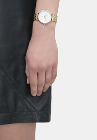 Goldfarbene Armbanduhr mit einem runden weißen Ziffernblatt und einfachen Stundenmarkierungen, kombiniert mit einem strukturierten goldenen Metallarmband. Die Hand ruht gegen ein schwarzes Lederbekleidungsstück.
