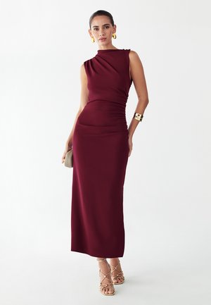 Žena má na sebe bezrukávové priliehavé bordové šaty s riasením, zlaté náušnice, náramok, drží béžovú kabelku a nosí zlaté sandále na vysokom opätku so remienkami.