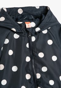 Marineblaue Regenjacke mit großen weißen Punkten, Reißverschluss und Kapuze. Der Stoff wirkt glatt und wasserabweisend.