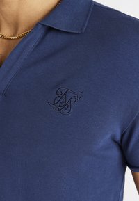 Lähikuva henkilöstä, joka pukeutuu tummansiniseen polo-paitaan, jossa on kirjailtu tumma logo rinnassa ja kultaketju kaulassa.