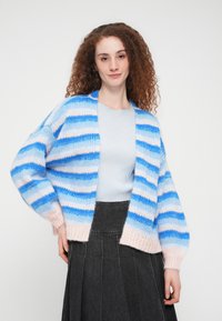 Ριγέ ζακέτα με διαφορετικές αποχρώσεις του μπλε και του ροζ, άνετη εφαρμογή, υφασμένη με υφή, φαρδιά μανίκια, φορεμένη πάνω από ανοιχτό μπλε τοπ και σκουρόχρωμη φούστα.