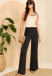 Črne široke hlače v kombinaciji s bež topom, ki ima črno obrobo. Model drži tkano torbo in nosi velike sončne očala.