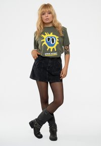 Olive green t-shirt with yellow and blue graphic print, paired with a black denim skirt and black knee-high boots with decorative buckles.