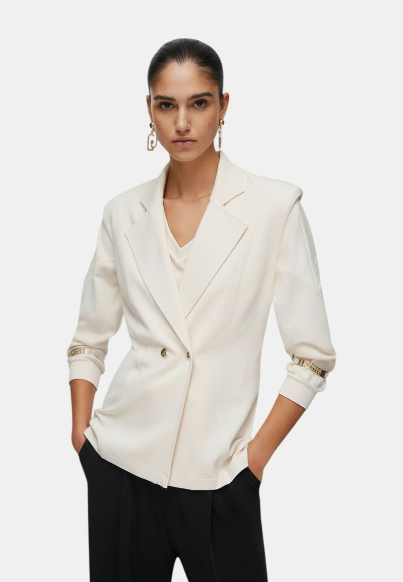 Mujer con un blazer blanco de doble botonadura y mangas remangadas, pantalones negros, manos en los bolsillos, y pendientes geométricos dorados.