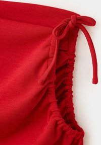 Gros plan d'un tissu rouge avec un bord froncé et plissé et un cordon attaché sur un vêtement, montrant la texture et les détails des coutures.