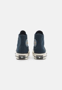 Blå canvas høye sneakers med en rund logo-patch, kontrasterende oransje detaljer og en hvit gummisåle med svarte striper.