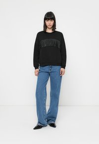 Zwarte sweatshirt met franjes over de borst, gecombineerd met lichtblauwe wijde spijkerbroek. Model staat tegen een witte achtergrond.
