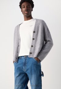 Grå ullcardigan med V-hals, ribbestrikkede mansjetter og fem brune knapper, kombinert med blå jeans og en hvit T-skjorte.
