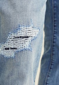Jeans de denim azul claro con un parche desgastado de textura tejida y bordes deshilachados en la rodilla, que muestran un diseño informal.