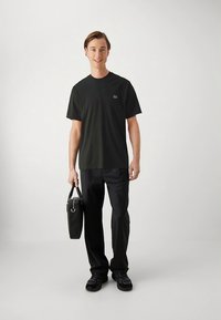 Svart t-shirt med rund hals och en diskret logotyp, kombinerad med svarta byxor och en svart väska. Modellen står självsäkert med avslappnade skor.