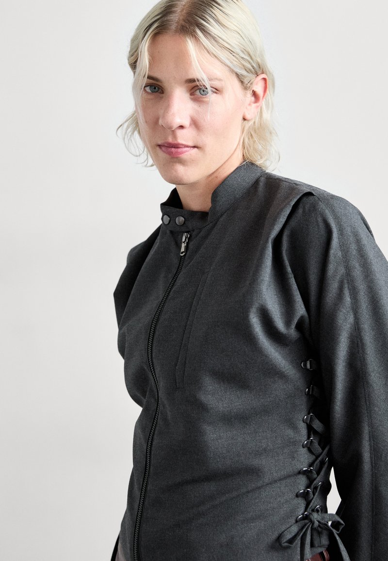 Giacca zip-up grigia scura con colletto alto, caratterizzata da dettagli di lacci laterali e una finitura liscia. Include una tasca frontale e bottoni a pressione.