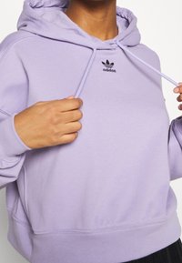 Helllila Kapuzenpullover mit Kordelzug und schwarzem Adidas-Logo auf der Brust. Verfügt über gerippte Bündchen und Saum für einen enganliegenden Sitz.