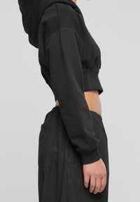 Svart kort hoodie med huva, ribbat avslut och långa ärmar, i kombination med löst sittande svarta byxor med sidofickor.