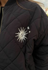 Fekete steppelt bomber dzseki, bal ujjon fehér spirál nap- és csillaghímzéssel. Texturált anyag, cipzáras záródással.