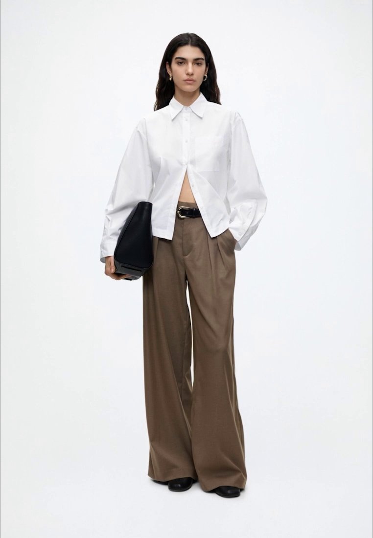 Femme portant une chemise blanche boutonnée, un pantalon large marron, une ceinture noire et des chaussures noires, tenant un grand sac à main noir sur fond blanc.