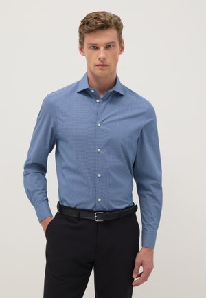 Jeune homme portant une chemise boutonnée bleue texturée ajustée avec un pantalon noir et une ceinture noire, debout devant un fond clair uni.