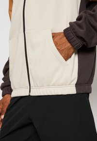 Chaqueta de hombre con cremallera en crema, marrón y gris oscuro. Cuenta con bolsillos laterales, puños elásticos y un dobladillo acanalado. Fabricada en una tela suave y texturizada.