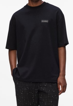 Mann trägt schwarzes Karl Lagerfeld T-Shirt mit Patch-Logo auf der Brust und strukturierte schwarz-weiße Hose, Hände in den Taschen.