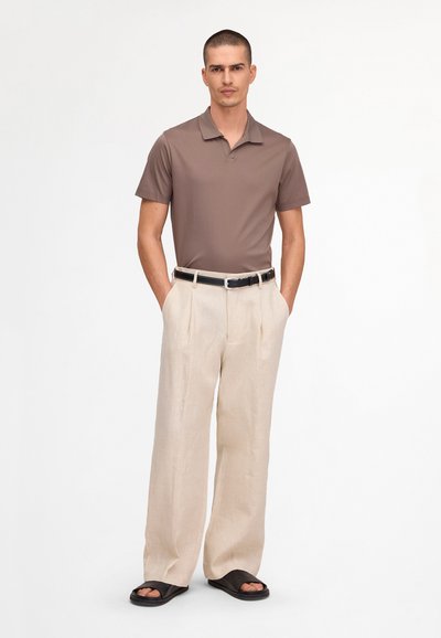 Homme debout, les mains dans les poches, portant un polo marron, un pantalon beige à jambes larges, une ceinture noire et des sandales noires slides sur fond blanc.