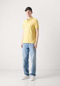 Gul kortermet polo skjorte, lys blå jeans, svart caps med hvit logo, og hvite sneakers. Enkelt design, avslappet passform, minimal detaljering.