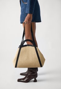 Beige gewebte Tragetasche mit schwarzen Ledergriffen, die eine strukturierte Form und eine strukturierte Oberfläche aufweist. Die Tasche wird in der Nähe von dunkelbraunen Knöchelstiefeln gehalten.