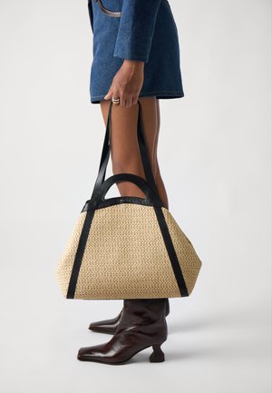 Borsa tote intrecciata beige con manici in pelle nera, caratterizzata da una forma strutturata e una superficie testurizzata. La borsa è posizionata vicino a stivali alla caviglia marrone scuro.