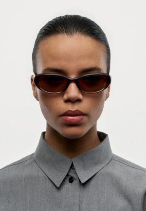 Persona con el cabello peinado hacia atrás, usando gafas de sol ovaladas oscuras y una camisa gris con cuello, mirando hacia adelante sobre un fondo liso.