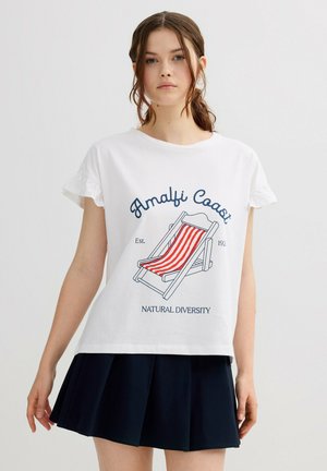 Joven mujer con camiseta blanca que lleva un gráfico de una silla de playa a rayas rojas y el texto "Costa de Amalfi, Diversidad Natural", combinada con una falda negra plisada.