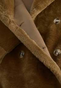 Brun shearlingsjakke med en glat, blød tekstur, der har et dobbeltradet design og store knapper, detaljeret med en kontrasterende inderfor.