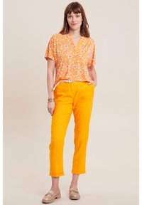 Sorange Bluse mit kurzen Ärmeln und weißem Blumenmuster, kombiniert mit leuchtend orangen Leinenhosen. Das Model trägt helle Loafers und ein Armband.