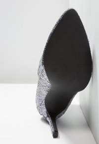 Chaussure à talon haut pointu avec semelle noire et dessus recouvert de sequins argentés, inclinée contre une surface blanche en angle.
