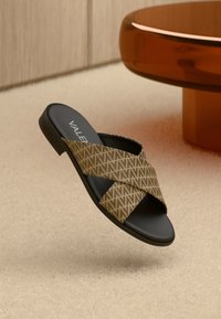 Brun mønstret slip-in sandal med kryssremdesign, svart såle og logo på innersålen. Avbildet mot en nøytral teksturert bakgrunn.