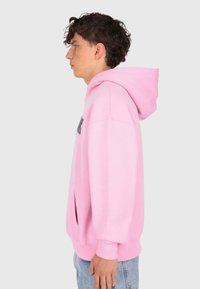 Junger Mann mit lockigem, dunklem Haar, der einen pinken Kapuzenpullover und helle Jeans trägt, seitlich stehend vor einem schlichten hellen Hintergrund.