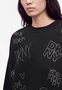 Femme portant un sweat-shirt noir avec des clous décoratifs formant des mots comme New York, DKNY et 1984 sur le devant.