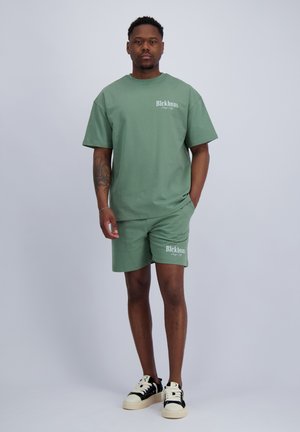 Man staat in een bijpassend groen oversized T-shirt en korte broek met witte tekstlogo's, zwart-witte sneakers, neutrale achtergrond.