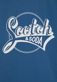 Blått stoff med et fremtredende hvitt grafisk design som har "Scotch & Soda" i stilisert tekst og en sirkulær aksent rundt.