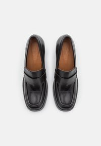 Black leather slip-on shoes with a smooth finish, rounded toe, and a decorative strap across the instep. Tan leather insole visible.