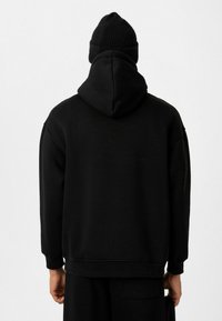 Zwarte hoodie met een relaxte pasvorm, textuurstof en geribbelde mouwen. Draag met een bijpassende zwarte beanie, van achteren bekeken.