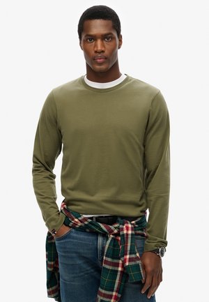 Młody mężczyzna ubrany w oliwkowozieloną koszulę z długim rękawem na białym t-shircie, niebieskie dżinsy, koszulę w kratę zawiązaną wokół talii i zegarek na nadgarstku, z rękami w kieszeniach.