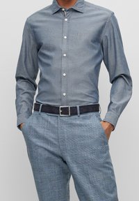Camicia azzurra chiusa con bottoni, colletto, maniche lunghe e bottoni bianchi, abbinata a pantaloni blu a quadri e cintura scura.